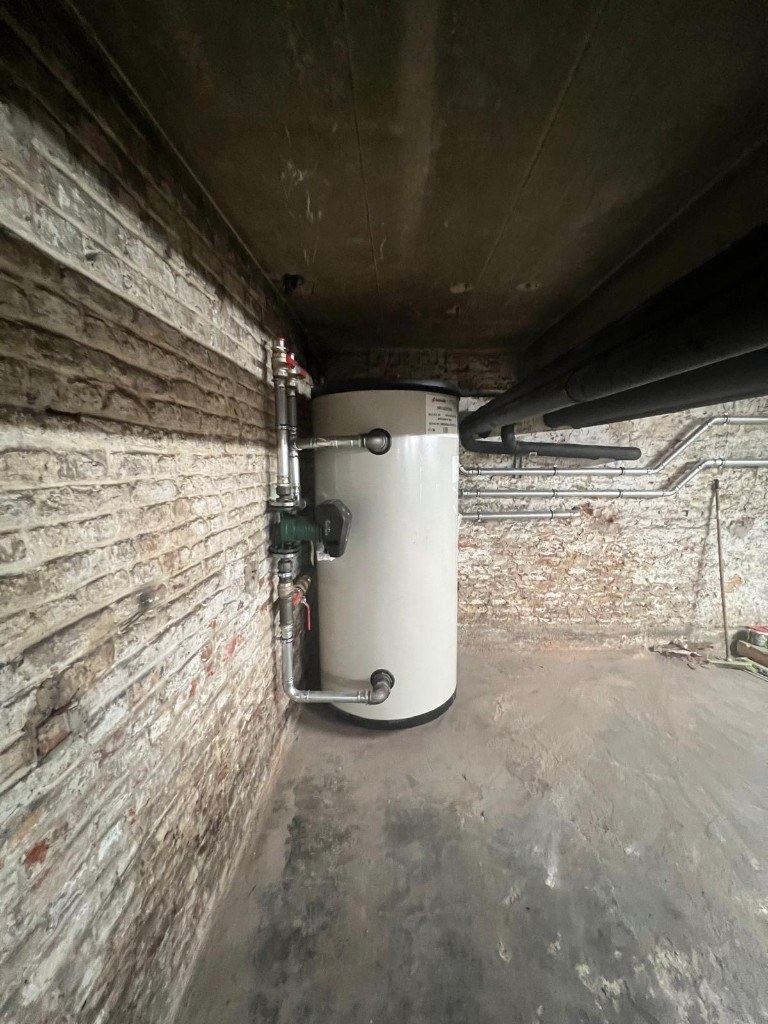 Chaufferie collective rénovée — ballon et tuyauterie en acier avec circulateur dans une cave en briques